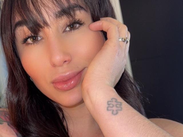 AgathaRuiz with brown hair, bangs, and glossy pink lips, holding her cheek with a hand showing a cross tattoo, wearing a silver ring. Her eyes are brown and slightly upturned.