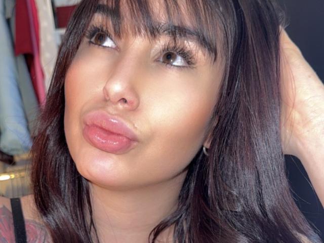 AgathaRuiz with pouty pink lips, brown eyes looking up, dark brown straight hair with bangs, slightly glossy skin, hand in hair, wearing black top, tattoo partially visible.