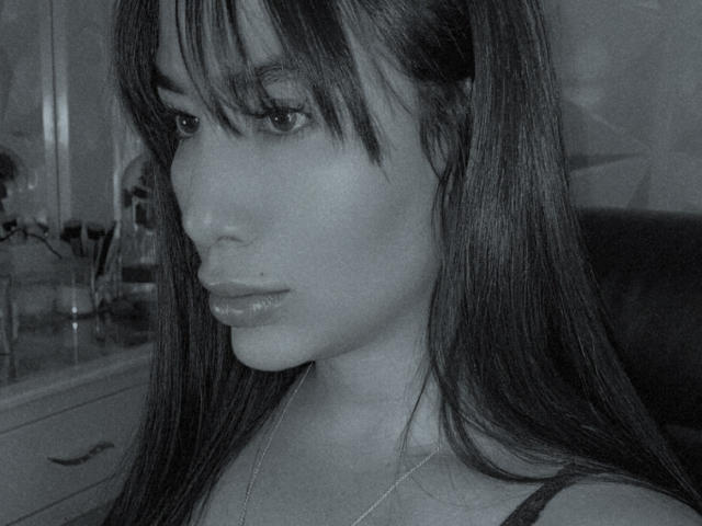Grayscale photo of AgathaRuiz with long straight hair, bangs, and a small beauty mark below her right eye. She's wearing a thin necklace, looking serious with slightly parted lips.