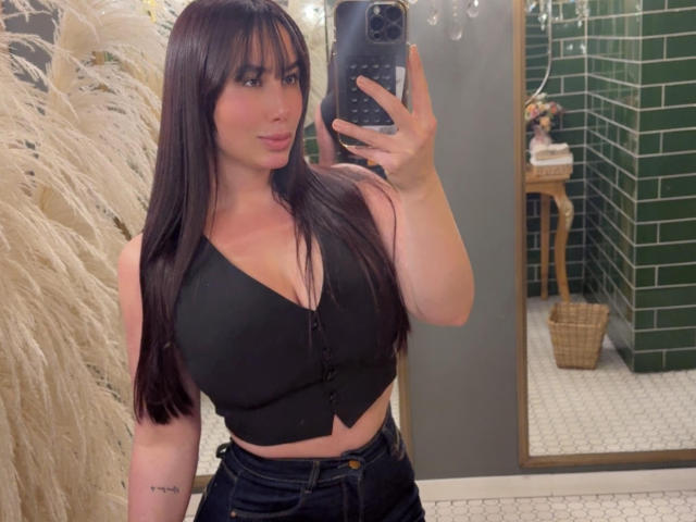 AgathaRuiz with long brown hair, wearing a black button-up crop top, showing her large tits in dark jeans, taking a mirror selfie, holding a black phone.