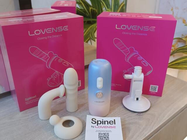 Lovense products: white curved butt plug, white cylindrical dildo, white suction toy, blue and white rechargeable vibrator, pink boxes, white product label, "SpineL by Lovense" in front.