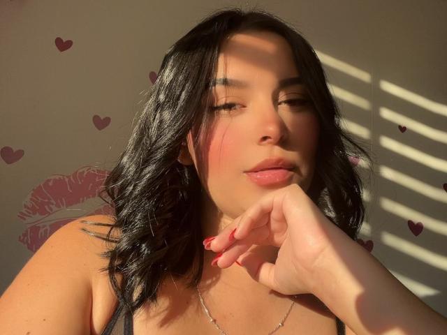 MiaAylen has black wavy hair, pink cheeks, and red nails, with sunlight casting shadows on her face, wearing a black top, and resting her chin on her hand.