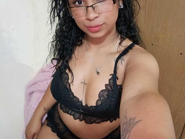 SexyStar69Lm with curly black hair, glasses, black lace bra, tattoos on chest and arm, holding phone, pink towel, brown door in background. Boobs, lace bra, medium skin.