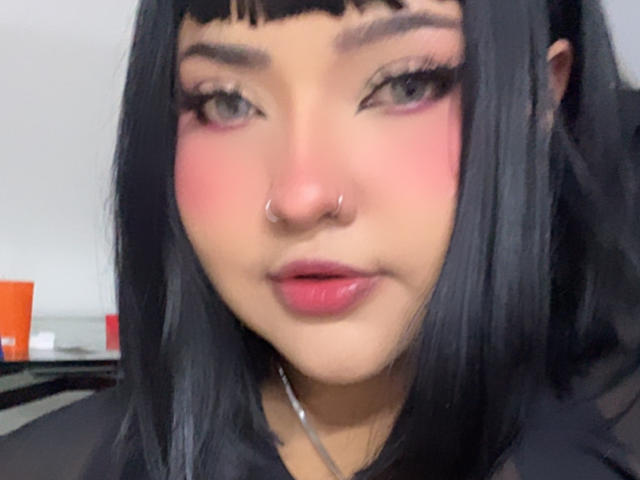 NathysWeety has black hair, pink blush, eyeliner, nose ring, and pink lips. She's looking at the camera with a slight pout, wearing a black top.