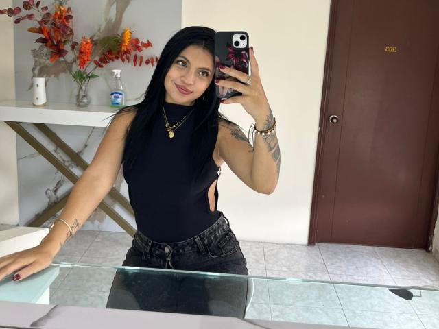 NyraVanel, com cabelo preto, vestindo uma blusa preta sem mangas, com recortes laterais, e calças jeans pretas, segurando um celular, sorrindo, com uma tatuagem no braço, em uma bancada de vidro, com uma porta vermelha..