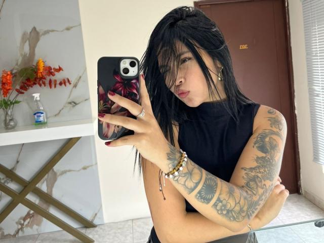 NyraVanel, avec des cheveux noirs, portant un haut noir sans manches, un bras tatoué, des ongles rouges, prenant un selfie, une porte marron en arrière-plan, une table en verre avec des fleurs orange et du savon..
