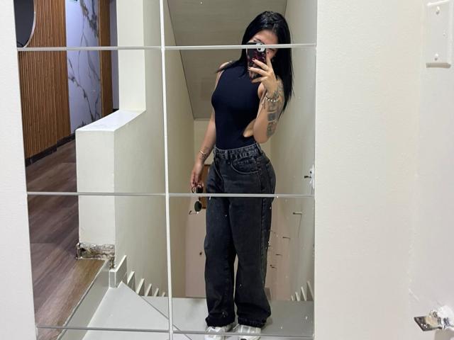 NyraVanel met zwart haar, die een strak zwart top draagt, donkere, hoge jeans, een telefoon vasthoudt, tatoeages op haar rechterarm, een spiegelzelfportret in een kamer met witte muren..