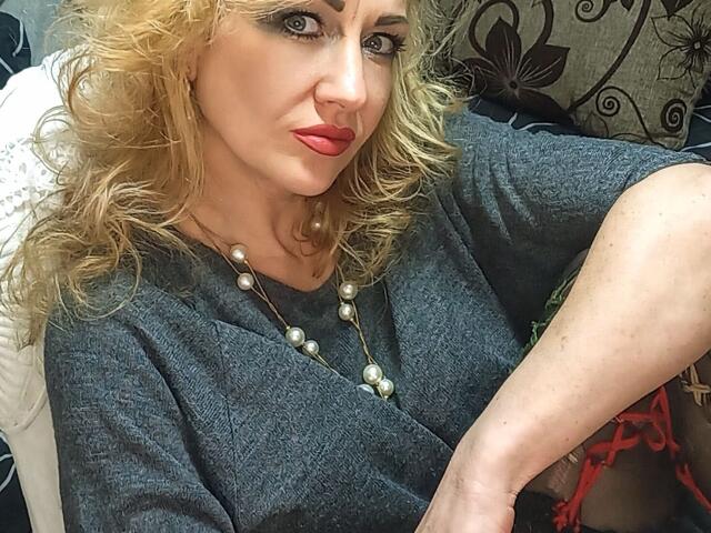 LilaniLoverX with curly blonde hair, red lipstick, gray tee, pearl necklace, and a slightly annoyed expression, sitting with crossed legs. Boobs are not visible.