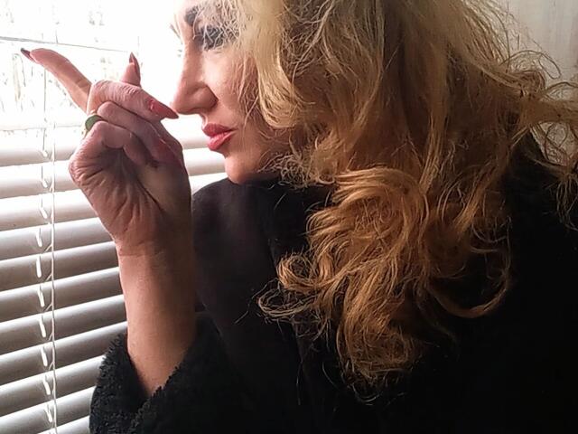 LilaniLoverX with wavy blonde hair, black dress, and red lipstick, points a finger near her nose, looking through white blinds. Her nails are painted red.