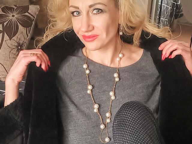 LilaniLoverX with blonde curls, red lipstick, and pearl necklace, wearing a gray top and black fur coat, hands on coat, red nails, and a ring, sitting on a couch.