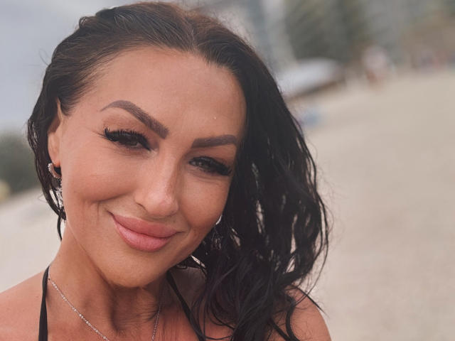 Close-up of AfriDetta with wet, wavy black hair, smiling slightly. She has dark eyebrows, long eyelashes, and pink lipstick. Her skin is tan, and she's wearing small earrings. A black halter top is visible. Her expression is relaxed and confident. The background is blurred, focusing attention on AfriDetta's face. The image has a soft, natural light. Her makeup is subtle, enhancing her natural features. The texture of her hair is slightly glossy. She has a small nose ring on her left nostril.