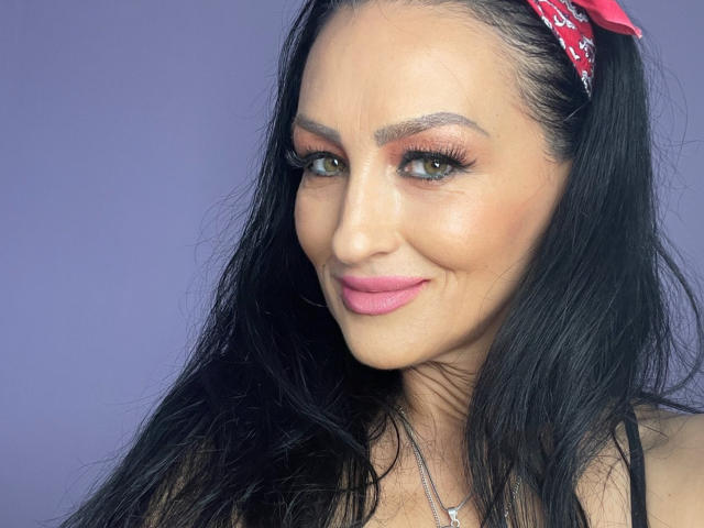 AfriDetta has long black hair, pink lipstick, and a red bandana. She's smiling with green eyes and dark eyebrows. Her tits are not visible, but she's cute.
