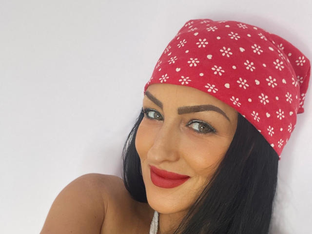 AfriDetta with dark hair, red lipstick, and a red floral-patterned headscarf, smiling at the camera. She has thick eyebrows and green eyes. Her tits are not visible.