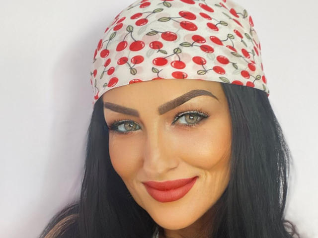 Close-up of AfriDetta with olive skin, green eyes, and dark eyebrows. She has long black hair, wears a red lipstick, and a white headscarf with a cherry pattern. AfriDetta's eyelids are lined with black mascara, and her eyebrows are thick and well-defined. Her smile is subtle, and her makeup is smooth and even. The headscarf's cherry pattern is bright red and green. The composition focuses on AfriDetta's face, with a simple, white background.