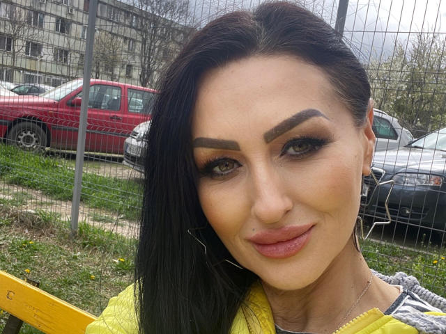 AfriDetta has long black hair, fair skin, and thick, dark eyebrows. She wears bold, dark eyeliner and long, curled eyelashes. Her lips are full and painted with a pinkish-brown lipstick. She smiles slightly, showing a small, pointed nose ring on her left nostril. She wears a yellow top with a gray pattern on the shoulder. The background includes a red car, a wire fence, and a building. The image is a close-up, focusing on her face.