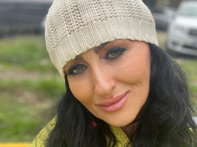 Close-up of AfriDetta with olive skin and black wavy hair. She wears a beige knitted beanie and a yellow-green sweater. Her green eyes have dark eyeliner, and she has pinkish-purple lipstick. The texture of the beanie is detailed with a ribbed pattern. AfriDetta's face is slightly turned to the right, showing a gentle smile. The background is blurred, featuring green grass and a white car. The image has a natural, slightly overcast lighting.