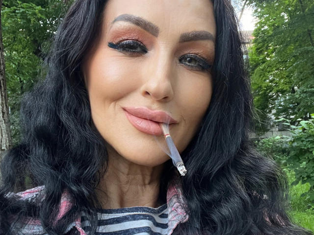Close-up of AfriDetta with long black wavy hair, light brown skin, and pink lipstick. She has thick, dark eyebrows and wears orange eyeshadow with black eyeliner. AfriDetta holds a cigarette in her right mouth corner. She wears a striped shirt under a pink and black flannel. Greenery is blurred in the background. The image focuses on her face, capturing a slight, confident smile. The texture of her hair is soft and wavy, contrasting with the smooth, matte finish of her lipstick. Her makeup is bold and eye-catching.