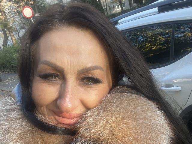 Close-up of AfriDetta with medium brown skin and dark, straight hair. She's wearing a furry coat, her brown eyes are accentuated with black eyeliner and long lashes. AfriDetta's eyebrows are thick and slightly raised. She's smiling with her lips closed. The background shows a silver car and some trees with yellow leaves. Natural light highlights her facial features. The image has a casual, real-life feel. AfriDetta's expression is confident and relaxed.