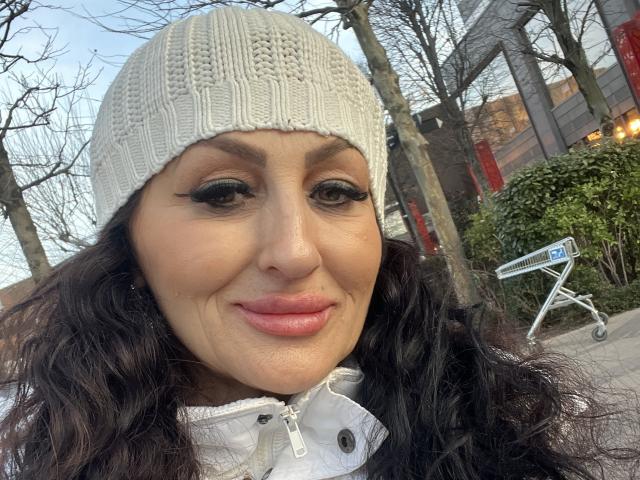 Close-up of AfriDetta with long black wavy hair, wearing a white knit beanie and a white winter coat with a zipper. She has dark, thick eyebrows, long black eyelashes, and pink lipstick. Her skin is light brown with visible freckles. She's smiling slightly. The background includes leafless trees, a green bush, and a street sign. The composition focuses on AfriDetta's face, with natural light highlighting her features. The image has a casual, outdoor setting.