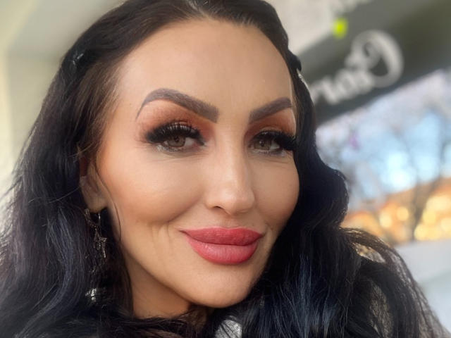 AfriDetta has dark wavy hair, thick eyebrows, orange eyeshadow, long eyelashes, and red lipstick. Her smile shows slight wrinkles on her upper lip.