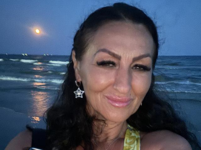 AfriDetta has a medium skin tone, dark brown wavy hair, and is smiling slightly. She wears a yellow halter top and star-shaped earrings. Her makeup includes dark eyeliner and pink lipstick. The blue evening light reflects on the wet beach in the background. The moon is visible in the dark sky. Her expression is relaxed and subtle. The image is a close-up.