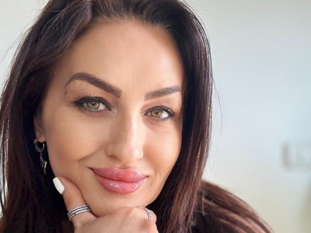 Close-up of AfriDetta with a confident, slight smile. She has medium-length, dark brown hair and is looking directly at the camera with green eyes. Her eyebrows are thick and arched, and she has full pink lips. She's resting her chin on her right hand, which has a silver ring on her index finger. Her skin is fair, and she's wearing subtle makeup with a natural look. The image is sharply focused on her face, with a soft, light background that is slightly out of focus.