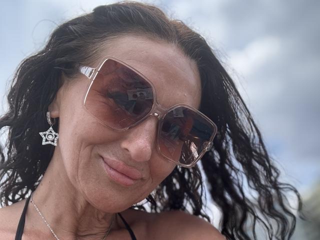 Close-up of AfriDetta with wavy, dark brown hair, wearing large brown-tinted sunglasses, a black halter top, and a star-shaped earring. She has a light brown skin tone and a subtle smile. The reflection in her sunglasses shows a blurred outdoor scene. The sky is cloudy, and the light is natural. Her facial features are sharp, with visible freckles. The image is sharp, with a focus on her face and upper shoulders.