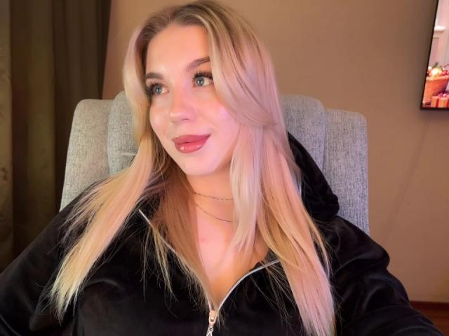 WildMaksi with blonde hair, blue eyes, pink lipstick, black zip-up hoodie, sitting in a gray chair, looking at camera, slight smile, necklace, TV on right side.
