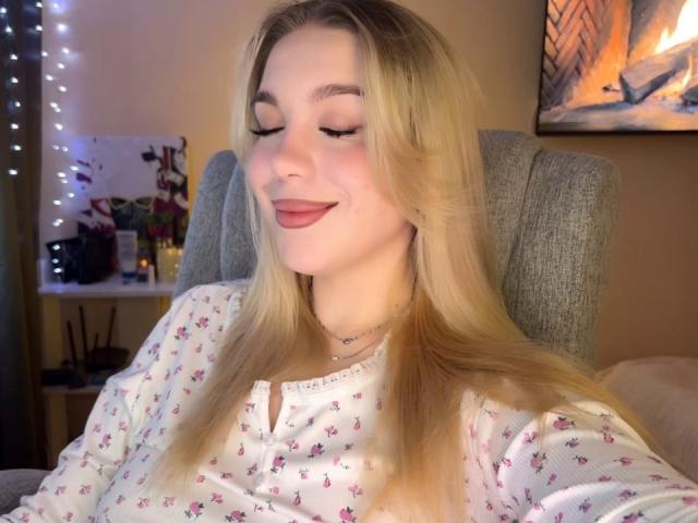 WildMaksi has blonde hair, closed eyes, and a pink lipstick smile, wearing a white floral shirt, sitting in a grey chair, with a necklace and fairy lights in the background.