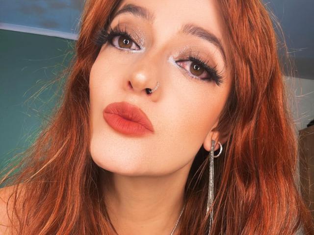 JaneRoa has red hair, brown eyes, and a nose ring. She's wearing bold eyeliner, long lashes, and red lipstick. Her silver earrings and parted lips are visible.