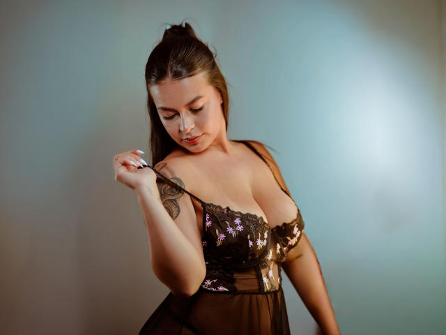 AgataVixen with big tits, wearing a sheer black lace dress, lifting a strap, brown hair in a high pony, tattoo on right shoulder, soft light, subtle smile.