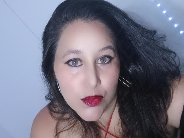 MadelineSade has dark, wavy hair, red lipstick, and black eyeliner. She's wearing a red top with a thin strap, and has silver hoop earrings. Her eyes are green.