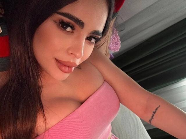 ArabianRoze with dark brown hair, full eyebrows, and long eyelashes, wearing a pink top that shows her big tits. She has a tattoo on her right arm and smiles.