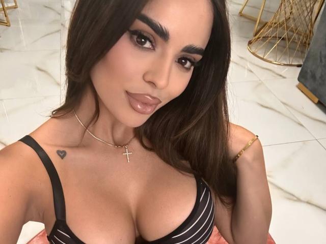 ArabianRoze with dark brown hair, full eyebrows, and glossy lips, wearing a black striped bra, showing her big tits, small heart tattoo, gold cross necklace, and gold bracelet.