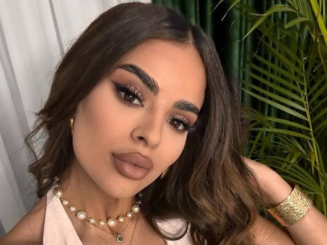 ArabianRoze has long, wavy brown hair, dark eyebrows, and thick, volumed eyelashes. She wears bold, matte brown lipstick and a pearl necklace with a small pendant. Her right arm has a gold bracelet. She has a light olive skin tone. The image is a close-up portrait with green leaves in the background. The composition highlights her face and makeup details. The lighting is even, emphasizing her facial features.