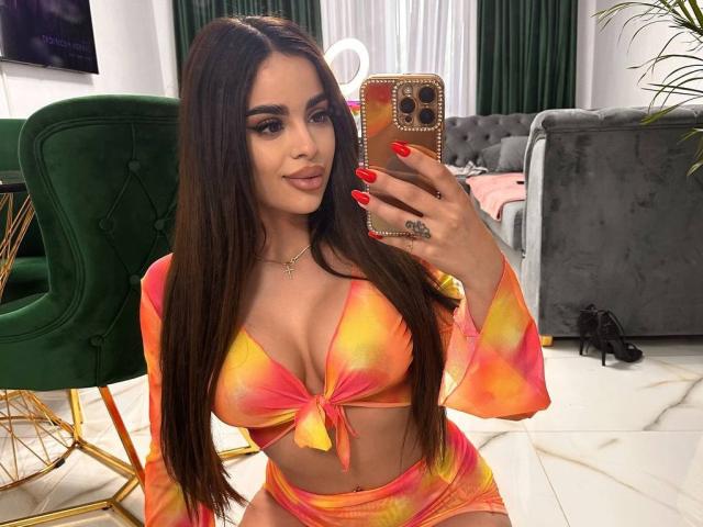 ArabianRoze con lunghi capelli castani, unghie rosse, un top corto arancione-giallo, pantaloncini abbinati, grandi seni, che tiene un telefono, sta scattando un selfie allo specchio, in salotto..