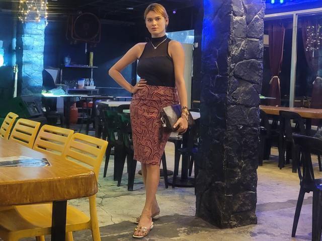 BeautyQueenTs stands with hand on hip, wearing black sleeveless top, red patterned skirt, silver sandals, holding black clutch, stone pillar behind, wooden table with yellow chairs.