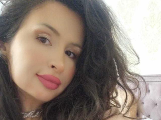 Close-up of JaneDivine with wavy, dark brown hair and a light skin tone. She has full pink lips and subtle brown eye makeup. Her gaze is slightly upward, with a gentle, confident expression. She wears a silver choker necklace. The background is blurred and light-colored. The image focuses on her face, highlighting her smooth skin and natural beauty. Slight shadow on the right side. Casual, intimate portrait style.