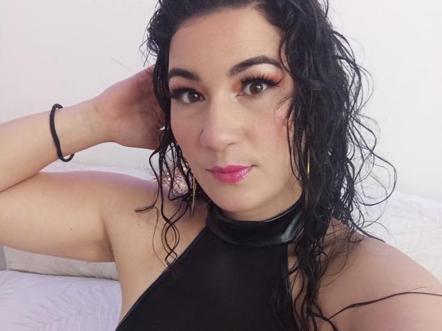 YessiSoto with wet black hair, wearing a black halter top, pink lipstick, and heavy eyeliner, looking at the camera with a slight smile. Her hand is on her head.