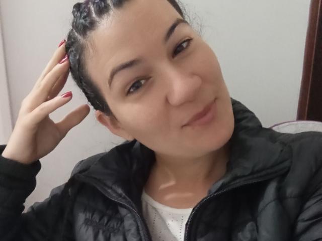 YessiSoto has dark braided hair, light skin, and a slight smile. She's wearing a black puffy jacket over a white shirt, with red nail polish, touching her head.