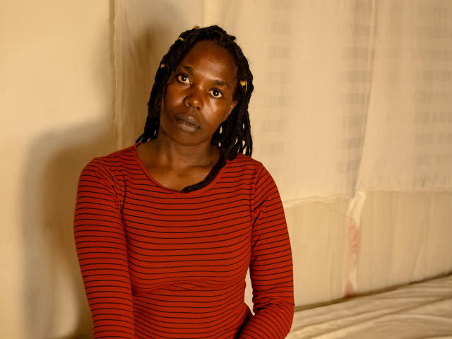 SweetApplex has dark brown skin, black dreadlocks, and is wearing a red striped long-sleeve shirt, sitting against a beige wall, looking serious with a slight frown.