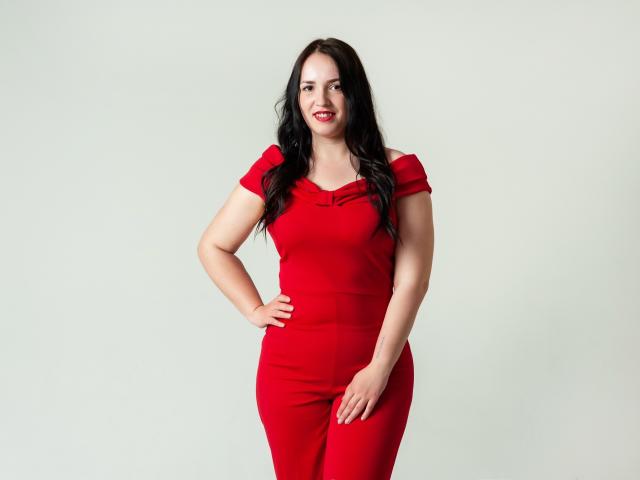 IsabelBlackberry stands against a plain background. She has long, wavy black hair and wears a bright red, short-sleeved dress that fits her curvy, medium-build figure. Her right hand rests on her hip while her left hand touches her thigh. She has fair skin and red lipstick, looking slightly to the left with a confident expression. The dress has a subtle cowl neckline, and the image is sharply focused with even lighting.
