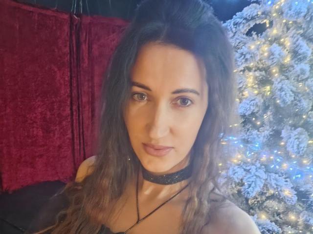 BanyReid has long, wavy brown hair and brown eyes. She wears a black choker necklace and a black top. Her expression is neutral, looking directly at the camera. In the background, there's a red velvet curtain on the left and a white, snow-covered Christmas tree with yellow and blue lights on the right. The image is slightly blurred, with a soft, warm light. The overall composition is centered on BanyReid, with the festive background adding contrast.