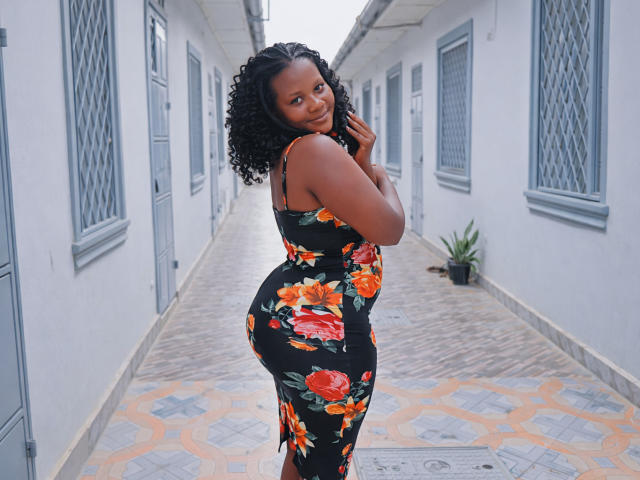 SexyBri with dark skin, curly black hair, wearing a black floral dress, looking back over her shoulder, showing her ass, standing in a narrow, gray-walled hallway.