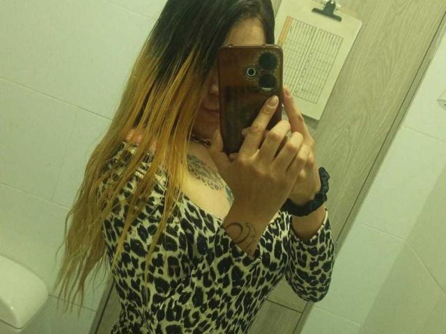 ErikaValeska with blonde to brown ombre hair, leopard print top, tattoos, taking a mirror selfie in a bathroom, phone covering her face, wrist scrunchie on her left hand.