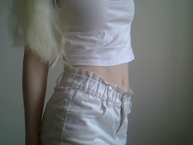 DitaVira stands in profile, wearing a white, slightly wrinkled crop top that reveals a flat, toned midriff. She has long, straight, platinum blonde hair that falls over her left shoulder. She wears high-waisted, shiny white satin shorts that have a slight sheen and a visible waistband. The image focuses on her torso and lower body, with a simple, unadorned background. The composition is minimalistic, emphasizing the textures of the clothing and her smooth skin.