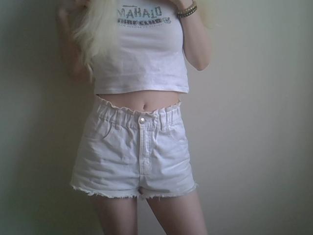 A photograph of DitaVira shows her from mid-thigh to just above her chest. She wears a white, cropped, sleeveless tank top with "Maid Princess Club" text and high-waisted, white denim shorts with frayed edges. Her long, blonde hair partially covers the top of the tank. She holds the tank top with her right hand, and a gold bracelet is on her right wrist. The image focuses on her midsection and lower body, with soft, natural lighting. The overall style is casual and minimalist.