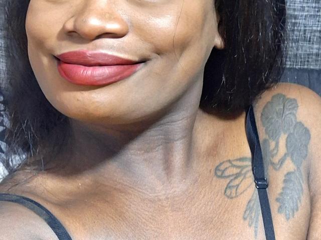 AlissaJordan's dark brown skin, red lipstick, and black shoulder tattoo on right side, with visible collarbone and black strap on left shoulder. Her face is slightly turned, smiling.