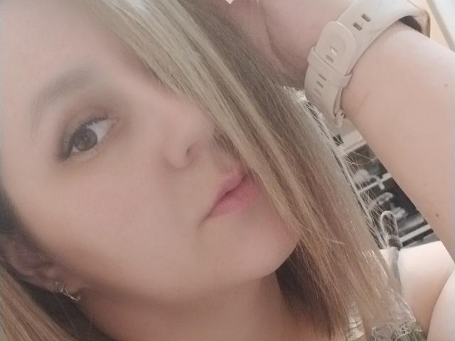 MabelMyriam's face, blonde hair covering one eye, pink lips, wearing a white smartwatch, slight pout, fair skin, brown eyes, small gold earrings, close-up selfie.