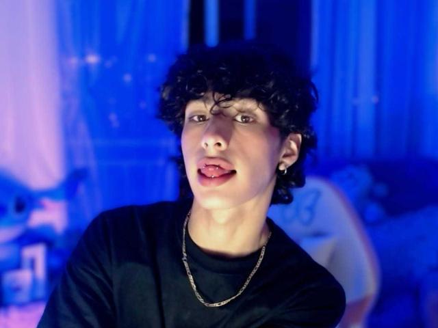SantiFolix has curly black hair, a black shirt, and a silver chain. SantiFolix's tongue is out, looking playful. SantiFolix has a small earring in their right ear.